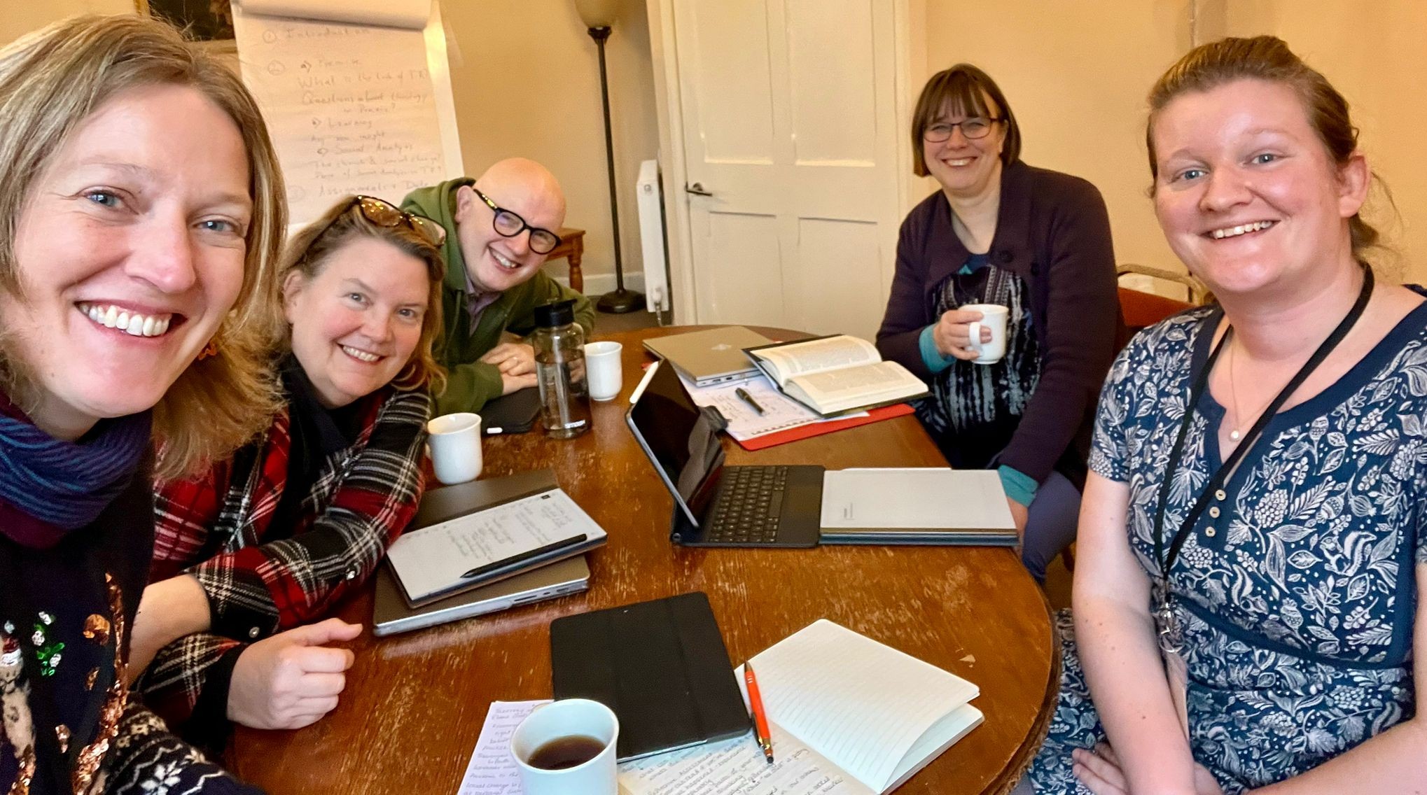 Five people are pictured sitting around a table, smiling. They have books and laptops in the middle of the table between them.