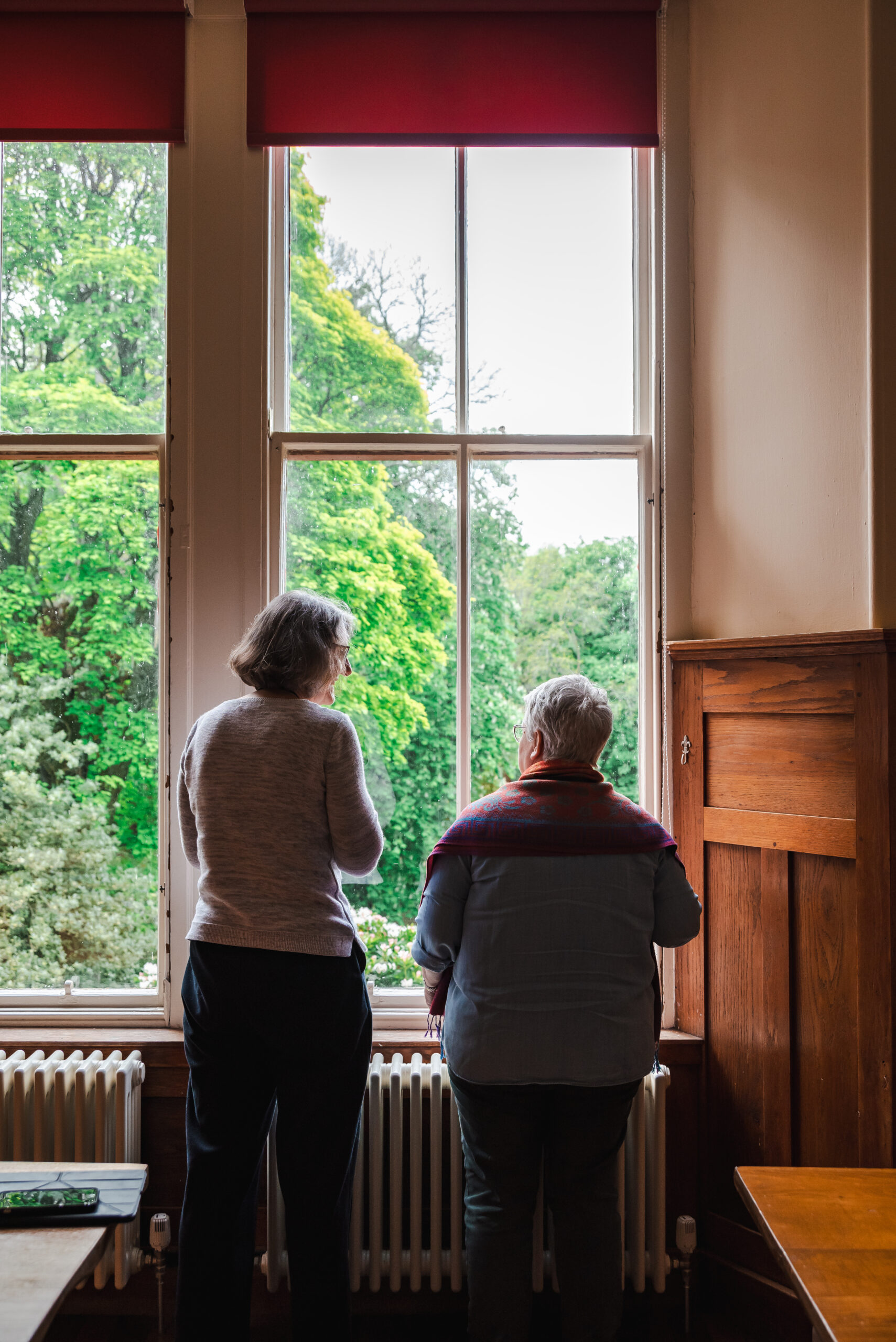 Two women (of varying heights) stand in the window, with their backs to the camera. The windows show lush green trees outside.