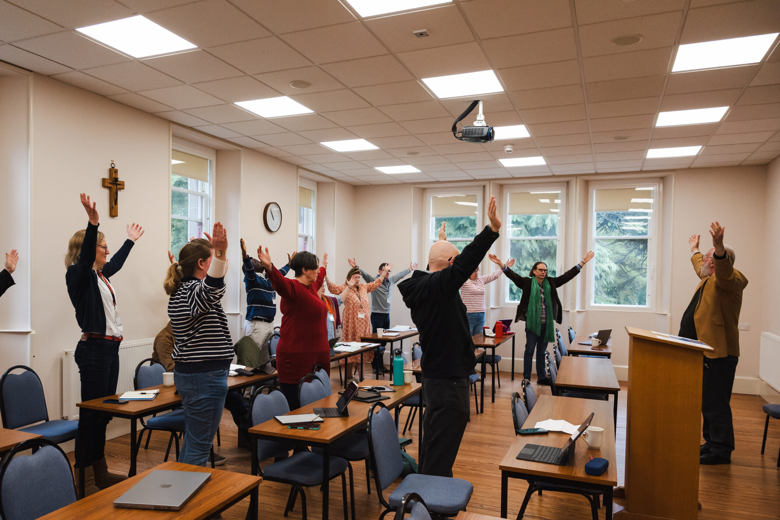 Approximately 20 students are standing in a classroom with their arms lifted up. A teacher stands at the front of the room.