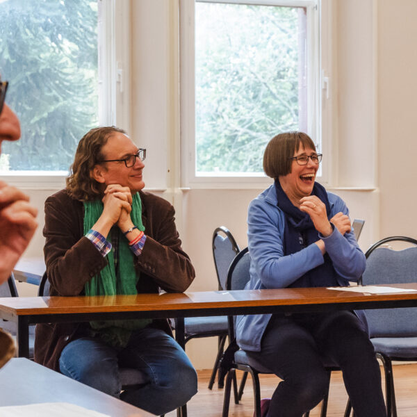 Two people sit at a a desk next to each other. They are smiling, or laughing and appear to be engaged with a wider context of discussion. Light is streaming through the windows behind them.