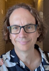 Picture of Daniel Farrell's face. Daniel has brown curly hair to his ears, white skin and is wearing glasses.