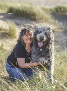 Carolyn smiling as she crouches next to her big, grey dog who has their tongue out. Sand and beach grass in the background.