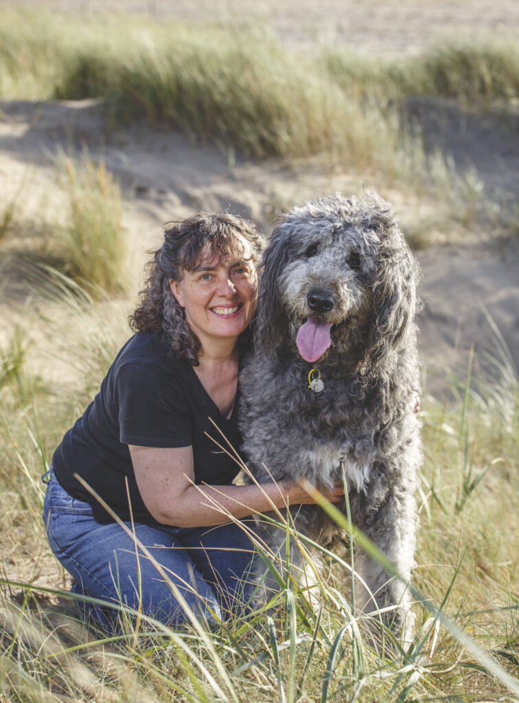 Carolyn smiling as she crouches next to her big, grey dog who has their tongue out. Sand and beach grass in the background.
